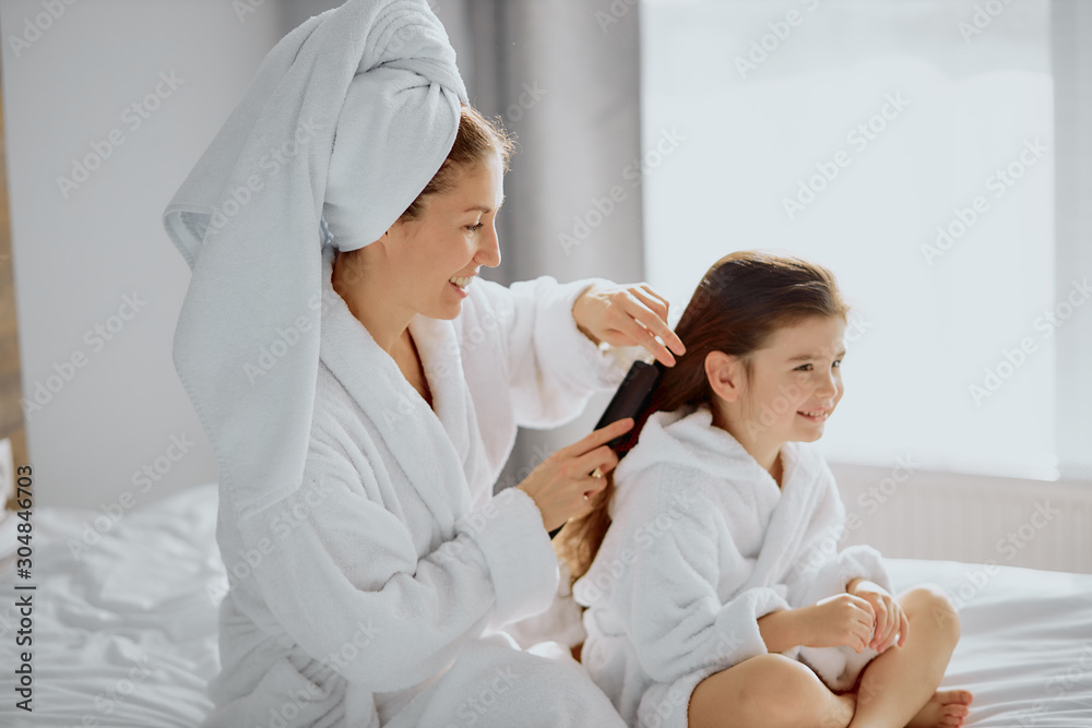 beautiful caucasian woman sit combing hair to lovely daughter, tidy up after shower in bright white bedroom. in bathrobes beautiful caucasian woman sit combing hair to lovely daughter, tidy up after shower in bright white bedroom. in bathrobes