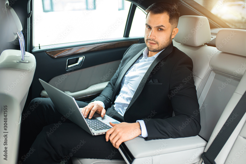 handsome young businessman sit with laptop while working in car, man looking at camera handsome young businessman sit with laptop while working in car, man looking at camera
