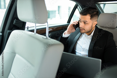 Handsome bearded man working on laptop while talking on phone in his luxurious car, side view © alfa27