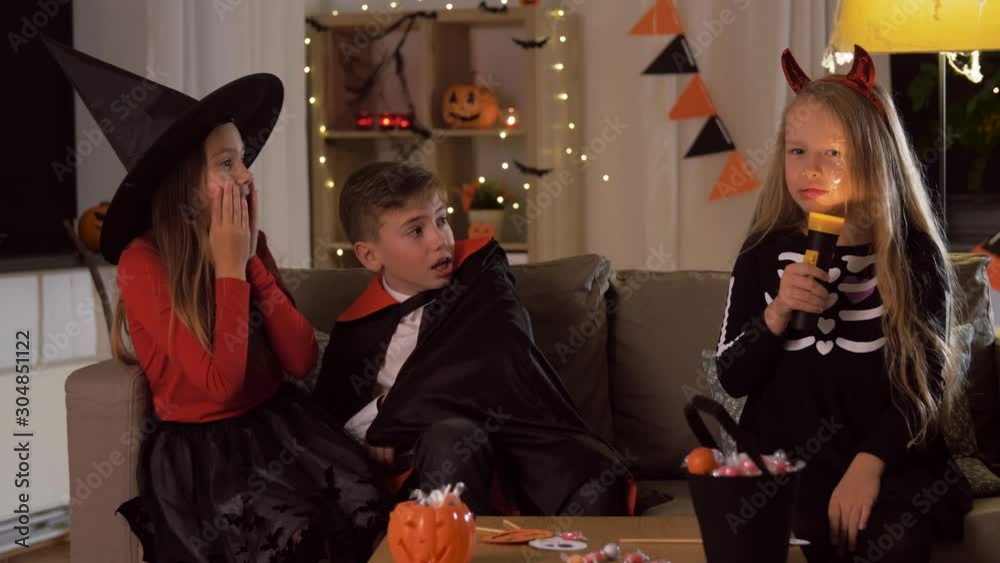halloween, holiday and childhood concept - smiling boy and girls in costumes playing with flashlight and scaring each other at home