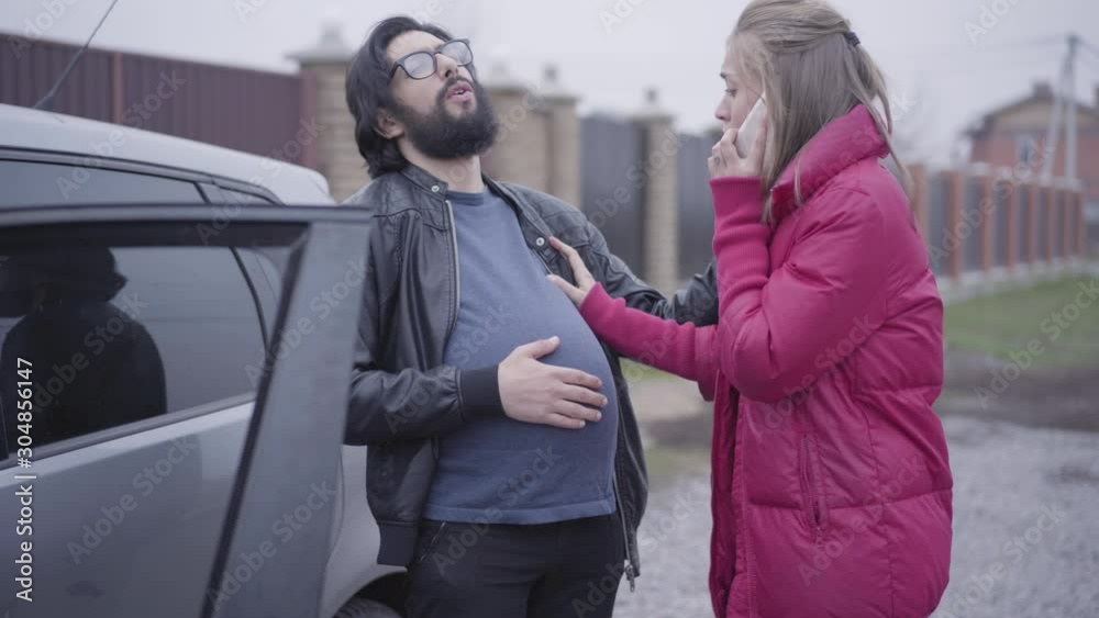 Pregnant Caucasian man in eyeglasses standing next to the car with his wife talking on the phone. Family exchanging their social roles. Joking, funny.
