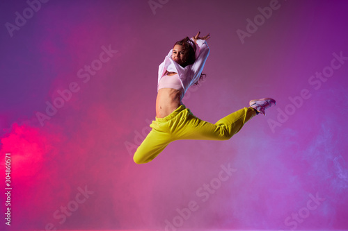 Smiling happy girl jumping high with raised hands, stretching legs intensively back, looking at camera with satisfied face, enjoying training in dance studio