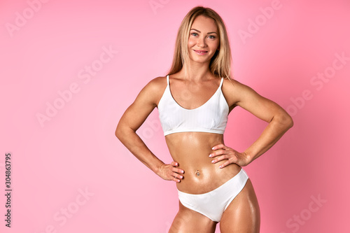Beautiful optimistic young woman with loose blonde hair and blue impressive eyes, looking at camera with cheerful expression, demonstrating sexy body in white swimsuit, keeping hands on hips, indoor © alfa27