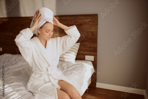Pretty attractive model sitting in comfortable room in hotel after bath procedures, wearing white soft bathrobe, wrapped head with clean bath towel, looking away, keeping hands on head, morning time