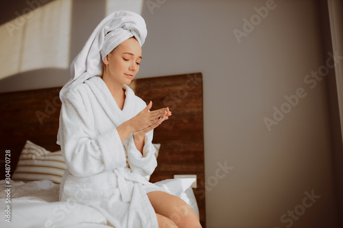 Charming young woman sitting on bed with depilatory sprips in hands, looking down, wants to do depilation after finishing bath procedures, wearing bathrobe and white towel wrapped on head