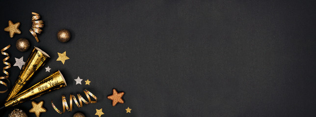 New Years Eve corner border banner of glittery gold stars, streamers, decorations and noisemakers. Top view on a black background.