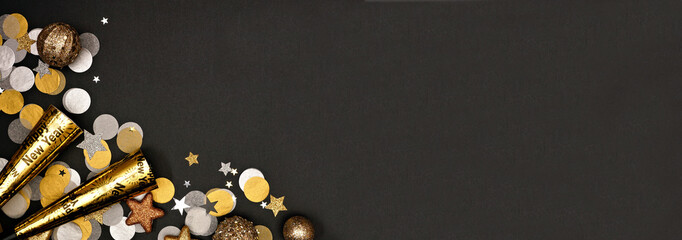 New Years Eve corner border banner of confetti, decorations and noisemakers. Overhead view on a black background.