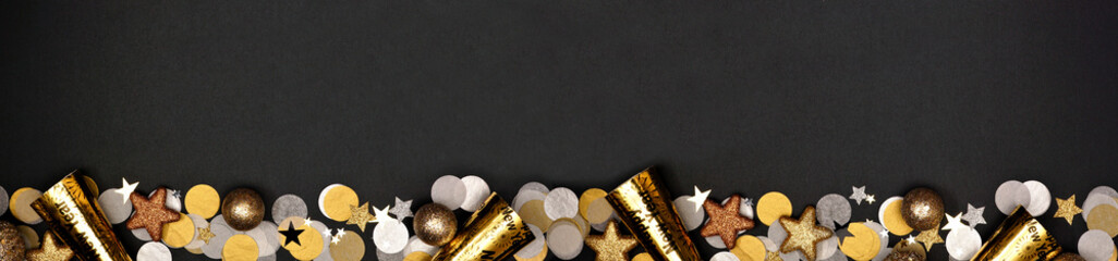 New Years Eve long border banner of confetti, decorations and noisemakers. Above view over a black background.