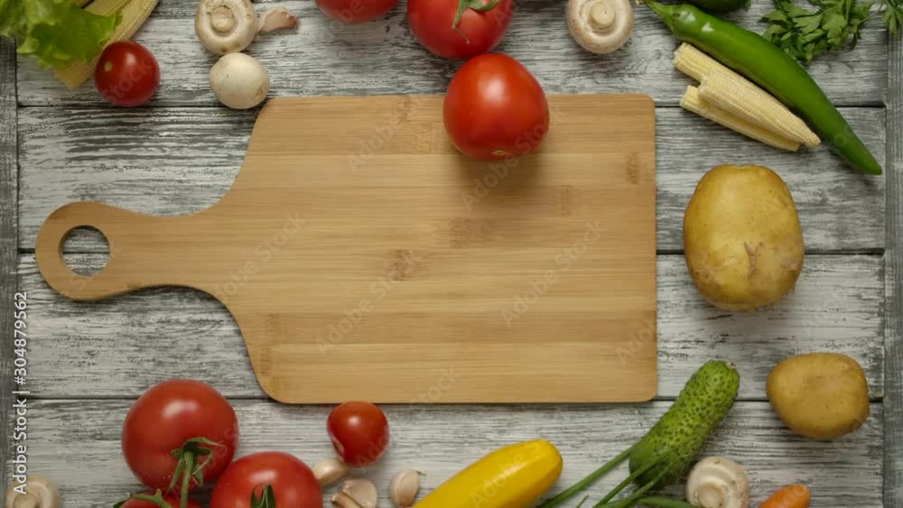 Male hand spinning red tomato on a wooden board. Top camera view from above over vegetarian products. Eggplant, cucumbers, potatoes, onions, paprika and mushrooms. Male hand spinning red tomato on a wooden board. Top camera view from above over vegetarian products. Eggplant, cucumbers, potatoes, onions, paprika and mushrooms.