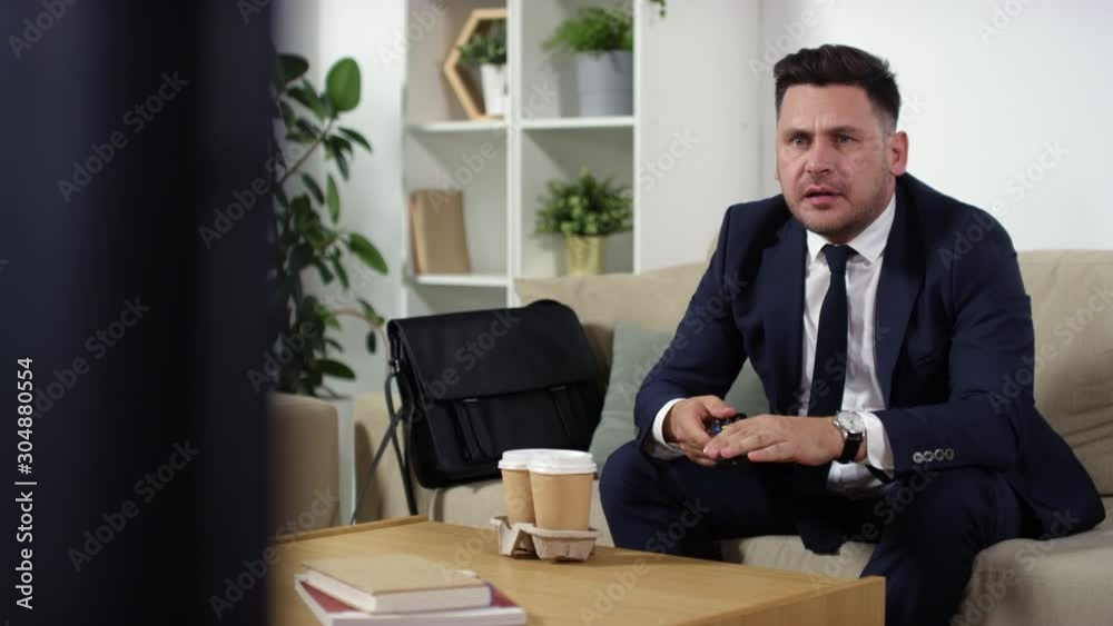 Nervous middle aged businessman in formal suit sitting on couch at home and watching sport match on TV. He getting upset, swearing and throwing remote control as his favorite team losing Nervous middle aged businessman in formal suit sitting on couch at home and watching sport match on TV. He getting upset, swearing and throwing remote control as his favorite team losing