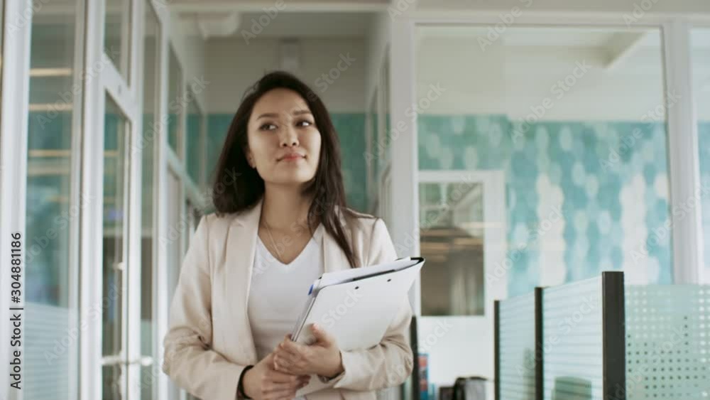 Attractive asian businesswoman holding documents and walking through the office towards the camera Attractive asian businesswoman holding documents and walking through the office towards the camera