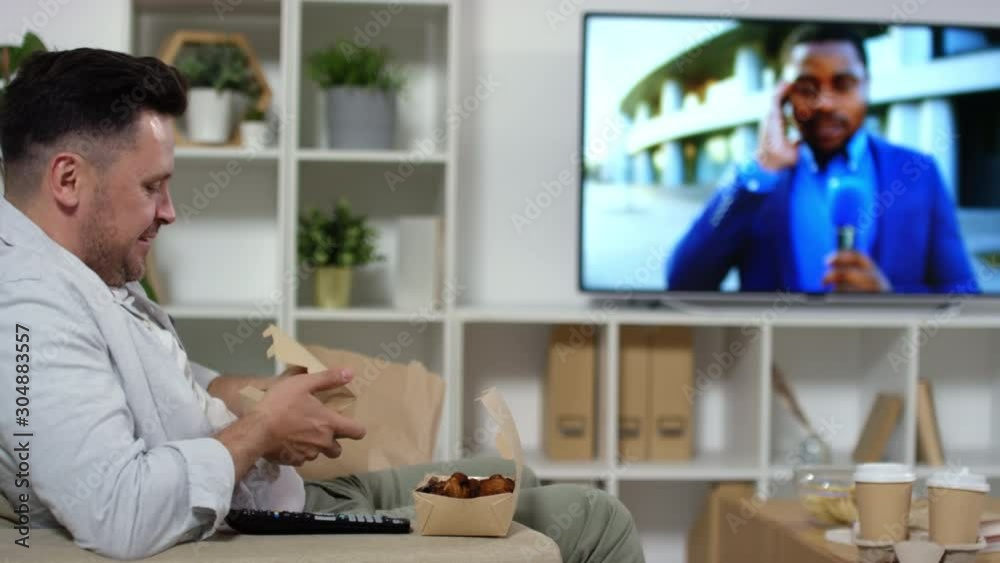 Mid-aged Caucasian man eating fries from takeaway box and switching TV channels with remote control while sitting in armchair and having meal in the living room at home