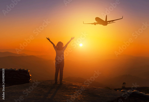 Silhouette back of woman stand raising up two arm celebrate enjoying on high cliff mountain while airplane flying in sunset time, freedom destination travel concept 