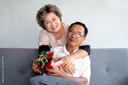 A asian senior man sitting on couch and hug by eldery woman - A wife give a red gift box to a husband for present in special day. Couple has smile and happy faces.