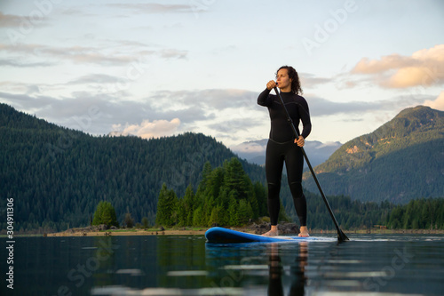 Adventurous Girl on a Paddle Board is paddling in a calm lake with mountains in the background during a colorful summer sunset. Taken in Stave Lake near Vancouver, BC, Canada.