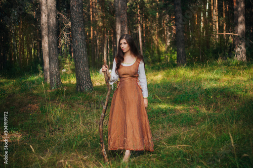 Young beautiful girl in medieval cowboy clothes, with a stick in hand. Barefoot on the ground. Against the background of the forest and green grass. A model with clean skin.