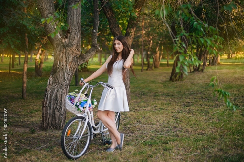 girl in a white skirt and a vest with a bicycle and a basket full of flowers in the park in the evening