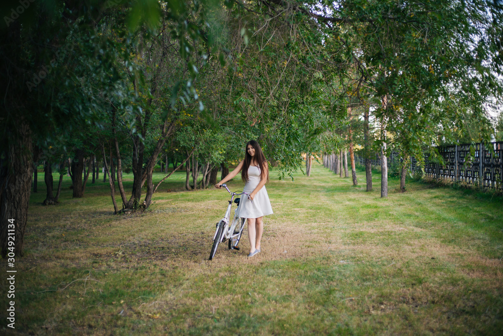 Fototapeta premium girl in a white skirt and a vest with a bicycle and a basket full of flowers in the park in the evening