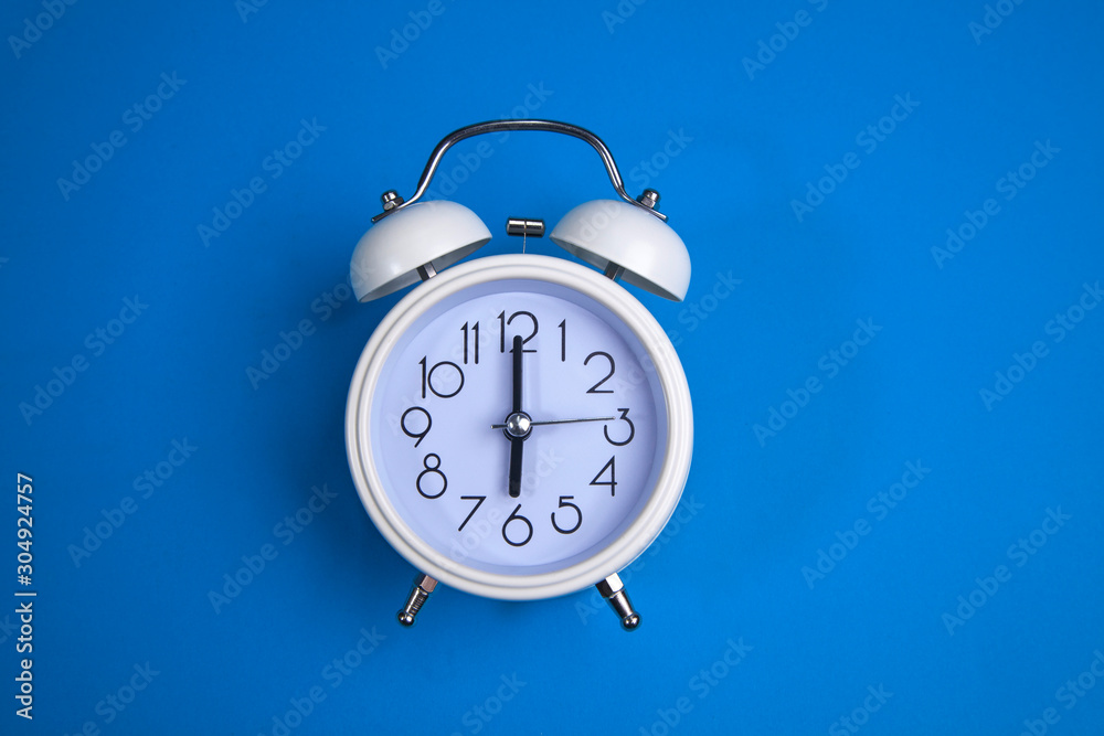 A white alarm clock on a blue background. a top view. a close-up picture representing 6 o'clock A white alarm clock on a blue background. a top view. a close-up picture representing 6 o'clock
