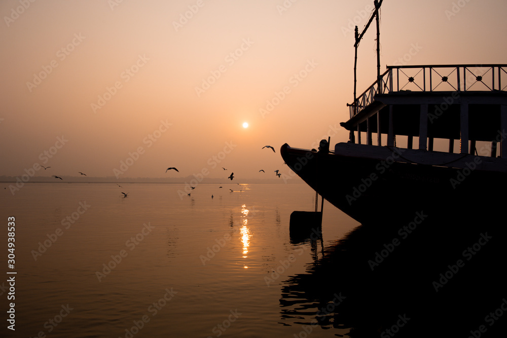 A hindu man praying during sunrise on the river Ganges in Varanasi, India. Fog, seagulls and boat silhouettes.