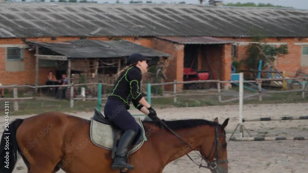 Horsewoman riding on brown horse and jumping the fence in sandy parkour riding arena. Competitive rider training jumping over obstacles in manege