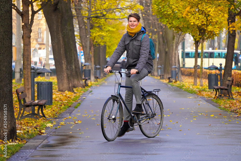 Obraz premium tall smiling guy of European appearance in glasses, with a yellow scarf and a gray jacket rides a Bicycle along the Avenue of the autumn Park. wet path after rain, fallen yellow leaves