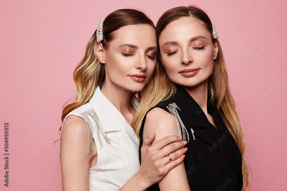 Fashion portrait of two beautiful young twins model girls with trendy makeup isolated at pink background. Beauty portrait of twins women with fashionable accessories, perfect makeup, and smooth skin