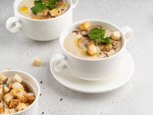 Delicious and creamy mushroom (champignon) cream soup in white ceramic bowls with green fresh parsley, flax seeds and crunchy croutons. White concrete background