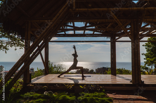 Yoga woman in bamboo studio. Woman doing yoga tree pose. Meditating and looking at beautiful sunset over Montanita beach town from wooden bamboo hut. Shot in yoga retreat in Ecuador. photo