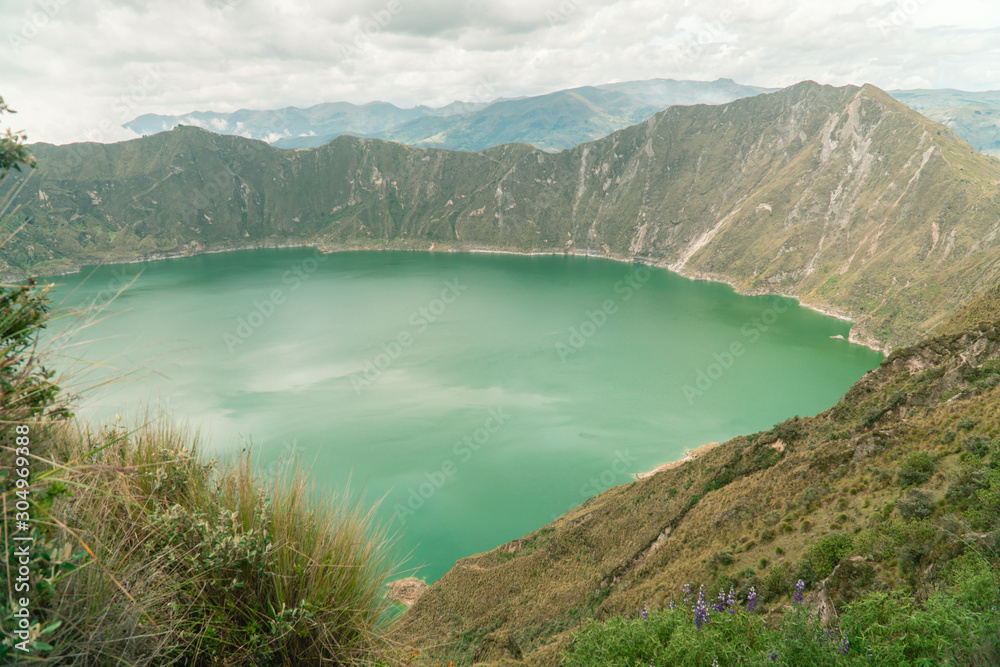 Volcano crater lake view, Quilotoa. Dramatic perspective of Quilotoa lake and volcano crater, with view of mountains, hiking path trail loop and cloudy sky from viewpoint. Shot in Ecuador. Green blue