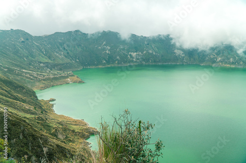 Volcano crater lake view, Quilotoa. Dramatic perspective of Quilotoa lake and volcano crater, with view of mountains, hiking path trail loop and cloudy sky from viewpoint. Shot in Ecuador. Green blue
