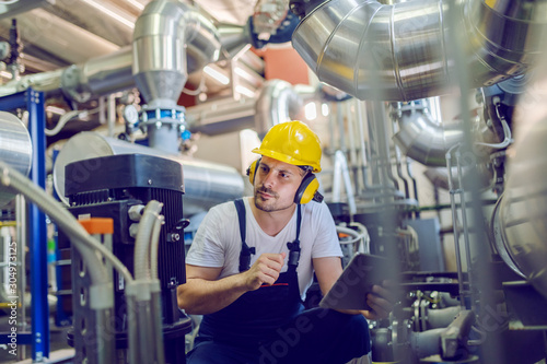 Serious caucasian handsome worker in overall, with hardhat and antiphons crouching and holding tablet while checking on motor. Factory interior.