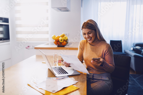 Cute caucasian smiling blonde woman in sweater holding bills in one hand and in other smart phone. On table are laptop and bills. Apartment interior. photo
