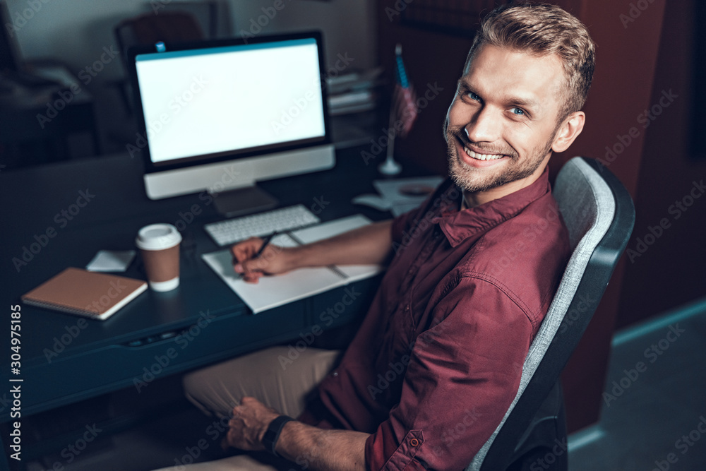 Happy young man is sitting at his office during late evening