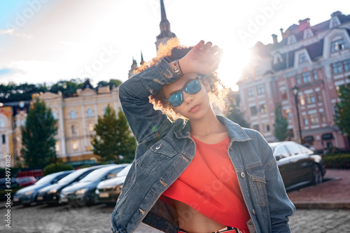 Outdoors Leisure. Young stylish african girl in denim jacket and sunglasses standing in sunlight touching head posing cool