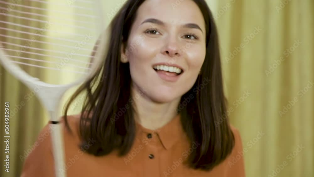 positive brunette girl in orange shirt dances with badminton racket having break in spacious coworking office slow motion