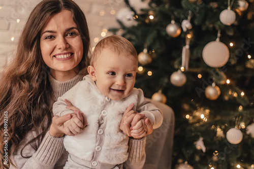 Happy family of mom with little baby hugging and celebrating new year in front of Christmas tree in decorated interior. Celebrating Christmas.