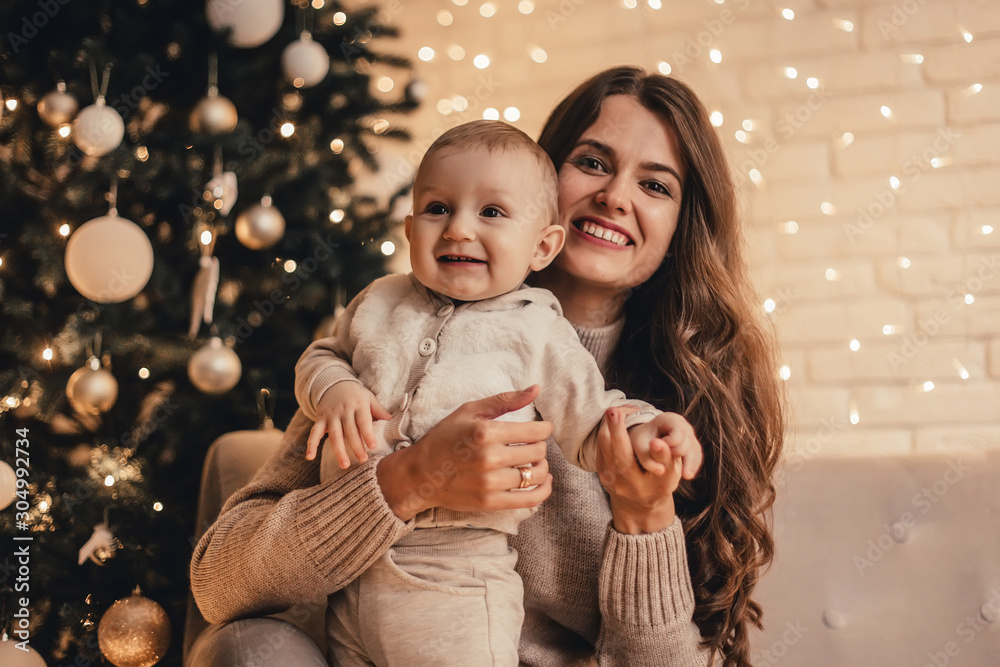 Happy family of mom with little baby hugging and celebrating new year in front of Christmas tree in decorated interior. Celebrating Christmas. Happy family of mom with little baby hugging and celebrating new year in front of Christmas tree in decorated interior. Celebrating Christmas.