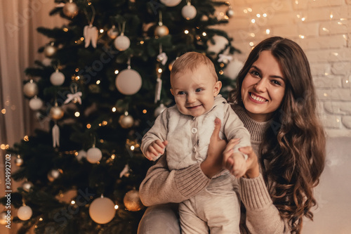 Happy family of mom with little baby hugging and celebrating new year in front of Christmas tree in decorated interior. Celebrating Christmas.