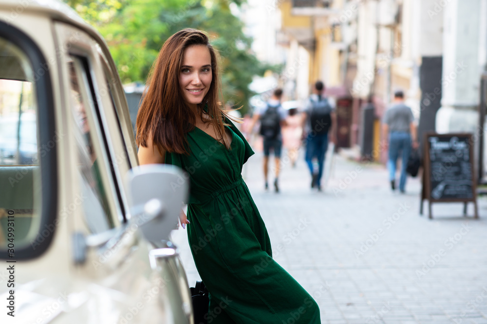 Fashion model wearing green overall posing outdoor next to Soviet vintage car. Young beautiful brunette caucasian woman walking summer streets. Beautiful girl, urban portrait.