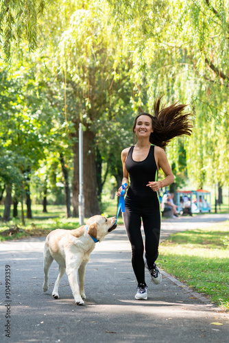 Cheerful pretty young woman walking and running with her dog in summer park. Active leisure, beautiful brunette girl at morning jogging with puppy