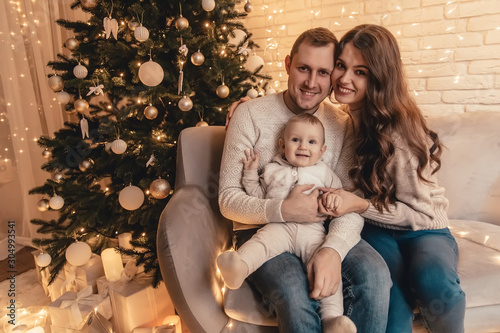 Happy family of mother, father and little baby boy hugging and celebrating new year in front of Christmas tree in decorated interior. Celebrating Christmas.