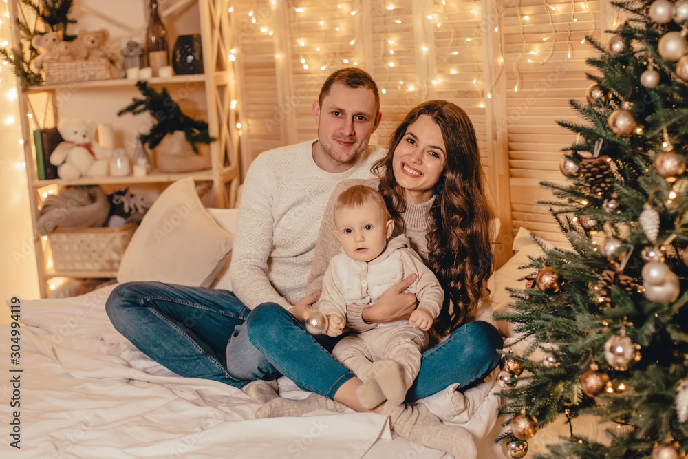 Happy family sitting on bed and celebrating new year near Christmas tree in decorated interior. Celebrating Christmas. Happy family sitting on bed and celebrating new year near Christmas tree in decorated interior. Celebrating Christmas.