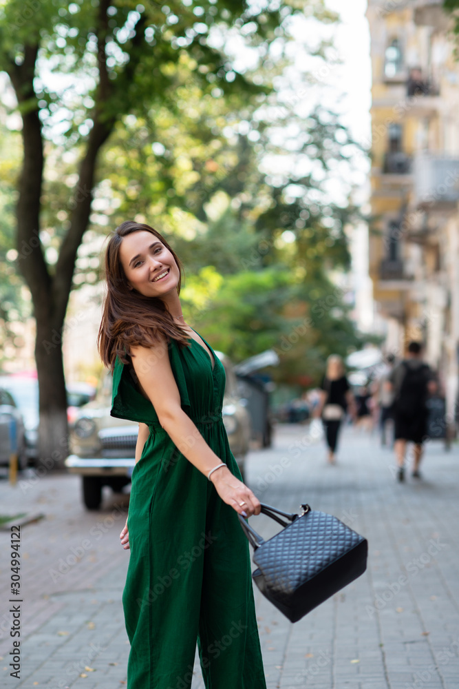 Fototapeta premium Fashion model wearing green overall posing outdoor. Young beautiful brunette caucasian woman walking summer streets. Beautiful girl, urban portrait.