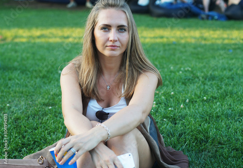 Beutiful young lady girl sits on grass in a park with smoothie or beverage in hand hangs out on a summer festival or party