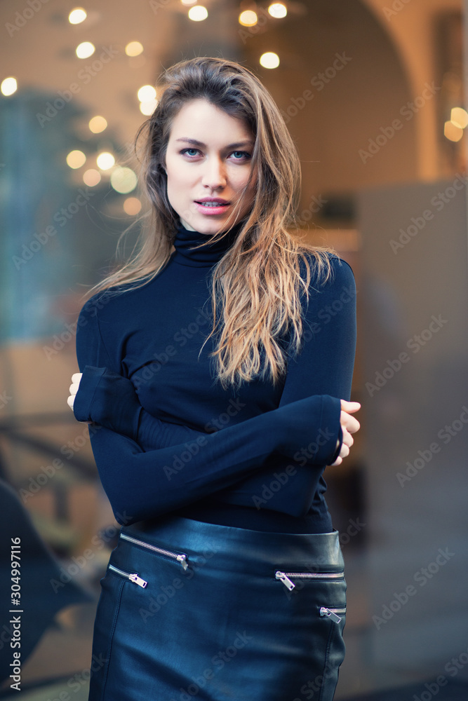 Fototapeta premium Young caucasian fashionable woman dressed in black clothes posing outdoor over Christmas Eve lights. Beautiful caucasian girl in Milan, Italy. Street fashion, winter holidays