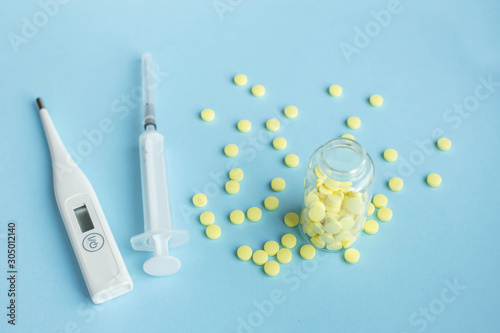 Yellow medical pills scattered from a glass jar, disposable syringe and thermometer on blue background with copy space for text. Healthcare and wellness concept. Top view, flat lay.
