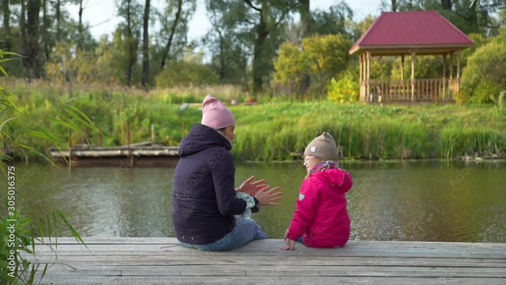 Little Girl and her Mother Talking while Sitting on a Pier near the Beautiful Lake in Autumn Day. Harmony with Nature, Leisure and People Concept Little Girl and her Mother Talking while Sitting on a Pier near the Beautiful Lake in Autumn Day. Harmony with Nature, Leisure and People Concept