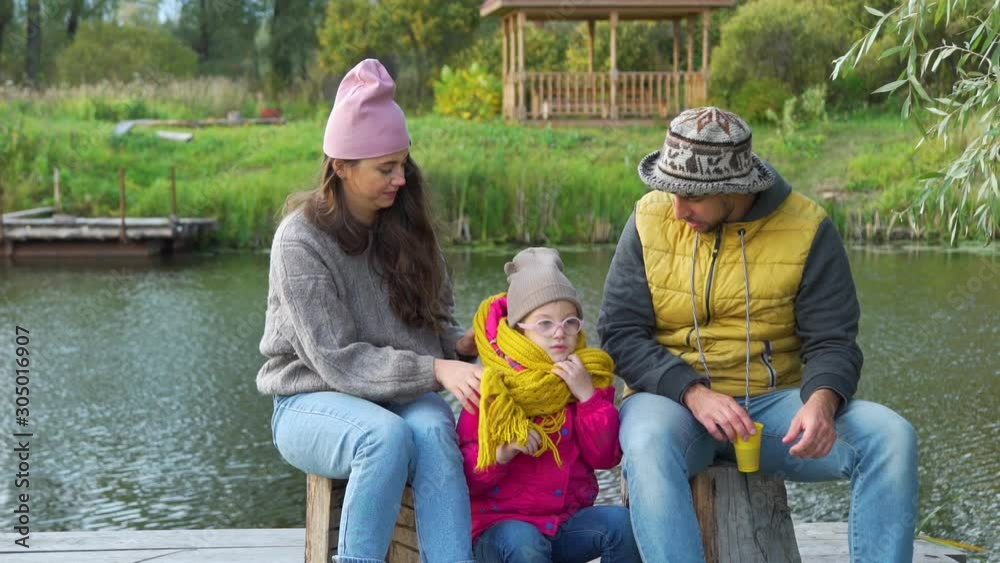 Young Family Having a Picnic near the Lake. Mother Helping her Daughter to Put a Warm Scarf. Family, Leisure and People Concept Young Family Having a Picnic near the Lake. Mother Helping her Daughter to Put a Warm Scarf. Family, Leisure and People Concept