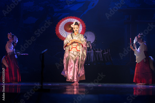 Traditional Japanese performance. Actress in traditional kimono and fox mark dancing with umbrella on a dark stage.